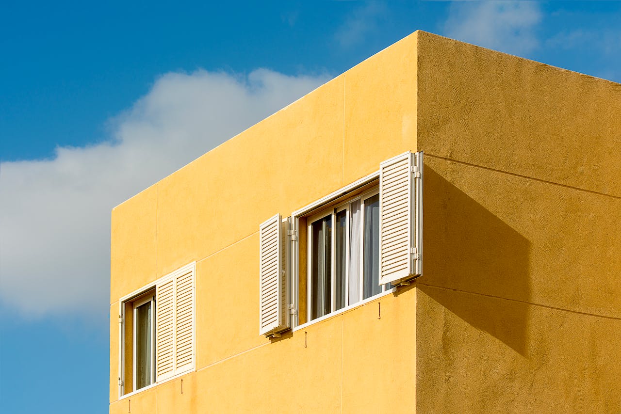 A modern yellow residential building facade with white shutters under a clear blue sky.