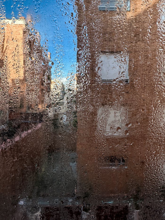 View through a rain-covered window showing urban buildings and clear blue sky in the background.